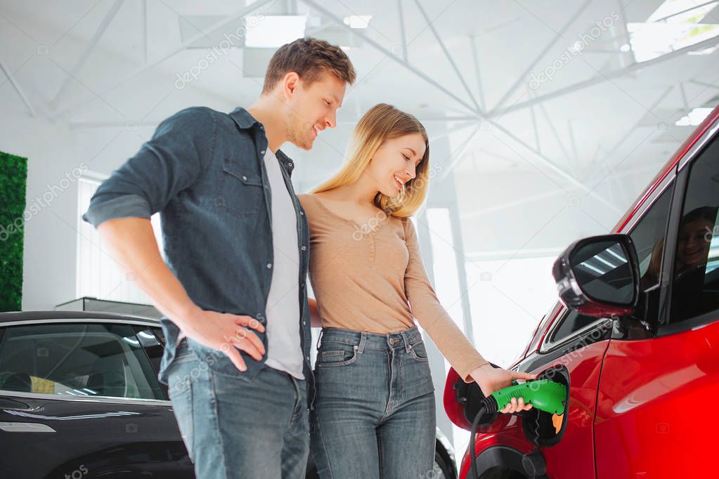 Young smiling couple buying first electric car in the showroom. Woman charging modern eco-friendly vehicle with the power cable supply plugged in