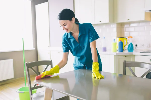 Pečlivý a příjemný vysavač stojí u stolu a vyčistěte jej. Nosí modré jednotné a žluté rukavice. Žena se dívá dolů. Pracuje v kuchyni. — Stock fotografie
