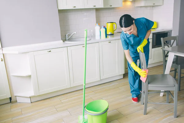 Concentrated and hard-working girl works in kitchen. She looks down and cleaning chair with orange rag. There are green bucket and mop on floor. — Stock Photo, Image