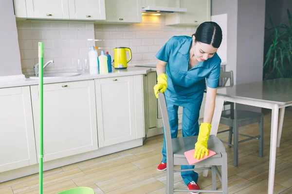 Kobieta Concent5rated kłania krzesło i oczyścić wilgotną szmatką. Pracuje w kuchni. Dziewczyna nosi niebieskie rękawice jednolite i żółty. — Zdjęcie stockowe