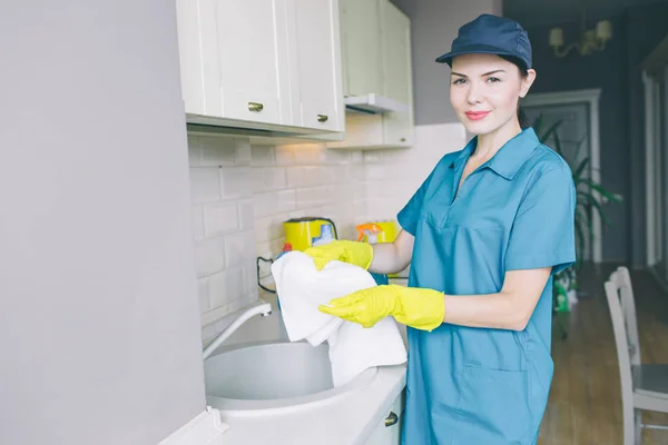 Nette Brünette steht am Waschbecken und hält ein weißes Handtuch in der Hand. Sie blickt zuversichtlich in die Kamera. Mädchen trägt blaue Mütze und Uniform. auch sie hat gelbe Handschuhe. — Stockfoto