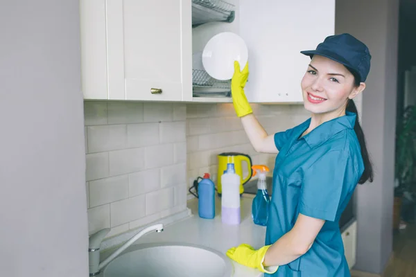 Positive and energetic girl stands at sink and look on camera. She smiles. Woman puts plate in cupboard. She looks careful. Girl is professional cleaner. — Stock Photo, Image