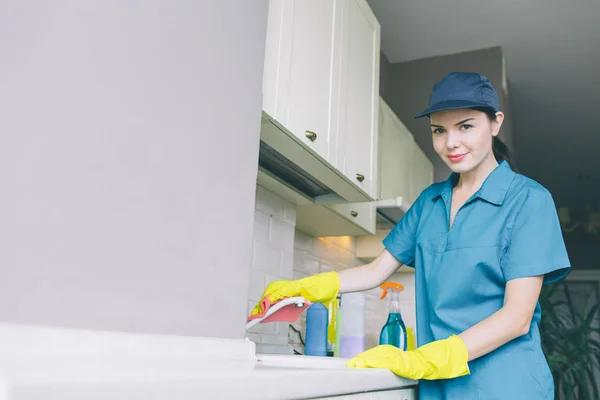 Ładne i atrakcyjne cleaner w cap czyszczenia płyn i wodę i spojrzeć na kamery. Ona nosi niebieskie rękawice jednolite i żółty. — Zdjęcie stockowe