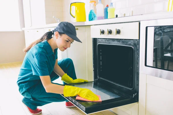 Hardworking woman sits in squad position and cleans door of stove. She is professional in cleaning. Girl wears gloves, cap and uniform.