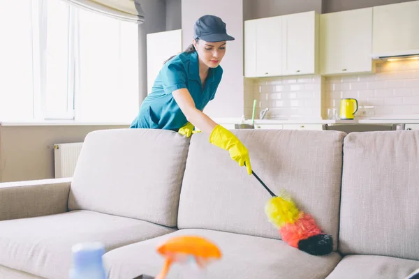 Ostrożny i skoncentrowany środek czyszczący działa w mieszkaniu. Ona używa kurz pędzlem na kanapie. Dziewczyna czyści delikatne. Ona jest osiągnięcie daleko kąty sofa. — Zdjęcie stockowe