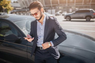 Yakışıklı ve meşgul bir genç adam siyah araba duruyor. O tutar ve telefon görünüyor. Dışarıda hava güneşli.
