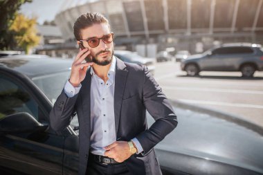 Ciddi ve yoğun genç işadamı arabaya standları ve telefonla konuşuyor. Güneş gözlüğü takıyor. Adam sağa doğru görünüyor. İyi giyimli.