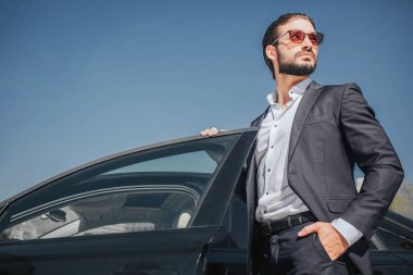 İyi giyimli işadamı resmini standı siyah arabadalar kapısını açtı. O tutan bir ve cebinde ve arabanın kapısını üzerinde bir tane. Genç adam pozlar.