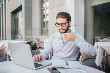 Konsantre adam masada oturur ve dizüstü bilgisayarda çalışır. Çay el üstünde tutun. Açık not defteri ve kalem masada vardır. İşadamı işleri.