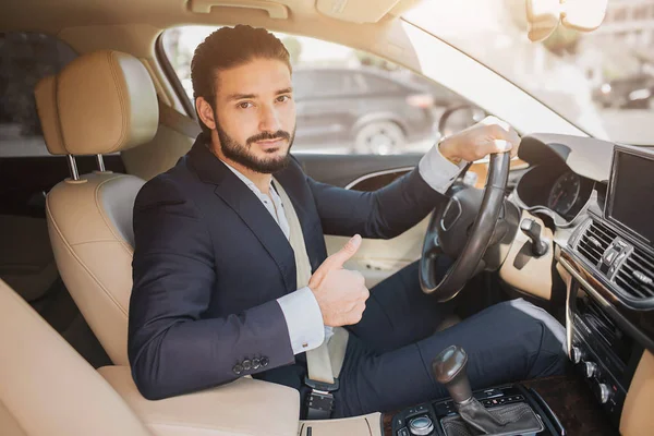 Милая фотография молодого бизнесмена, сидящего в роскошной машине и держащего большой палец вверх. Он держит одну руку на руле. Молодой человек рекомендует эту машину . — стоковое фото