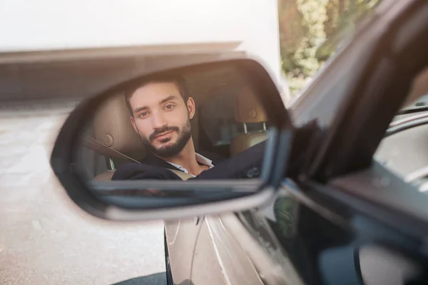 Фото человека, сидящего в машине и смотрящего на зеркало крыла. Он немного заикается. Молодой человек доволен своей машиной. На улице солнечно. . — стоковое фото
