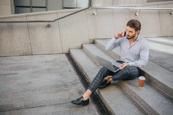 Picture of young businessman sits on steps and works. He talks on phone and looks at laptops screen. Young man keeps his legs crossed. — Stock Photo, Image