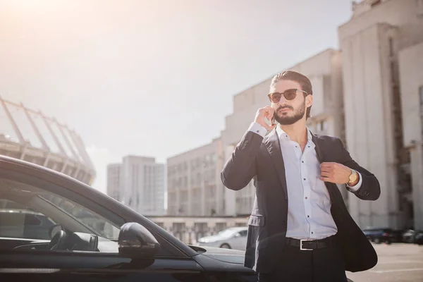Sérieux et bien habillé jeune homme debout à la voiture dans la suite et parle au téléphone. Il tient une partie de sa veste. Il porte des lunettes de soleil. Il fait beau et ensoleillé dehors . — Photo