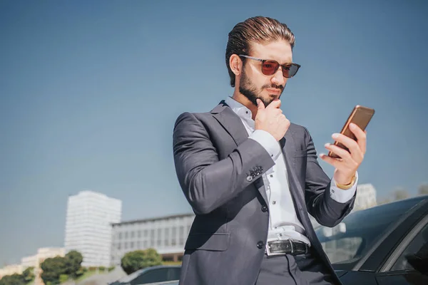 Un joven ocupado mira el teléfono que tiene en las manos. El tipo tiene otra mano en la barbilla. Él piensa. Joven se para en coche negro . — Foto de Stock