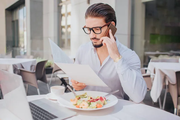 젊은 사업가 레스토랑에 전화에 회담 테이블에 앉는 다. 그는 노트북 화면에 보인다. 샐러드 그릇 및 테이블에서 커피 한잔. — 스톡 사진