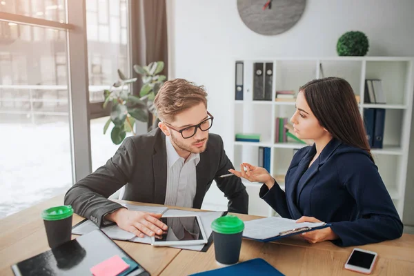 Un jeune homme concentré s'assoit ensemble avec une femme et travaille. Il regarde les papiers dans sa main. Elle explique à un mec. Travail productif . — Photo