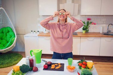 Vegan beautiful blonde woman has fun with cucumber while cooking raw vegetables in the kitchen. Raw food diet. Vegetarian food. Healthy eating clipart