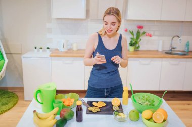 Vegan beautiful blonde woman with smartphone photographing organic orange in the kitchen. Healthy food. Diet detox clipart