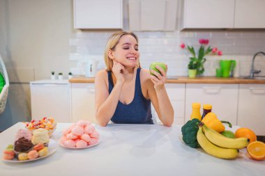 Young smiling woman with green apple in one hand choosing between healthy and unhealthy food in the kitchen. Difficult choice between fresh fruit vegetables or sweets. Dieting. Diet. Healthy Food clipart