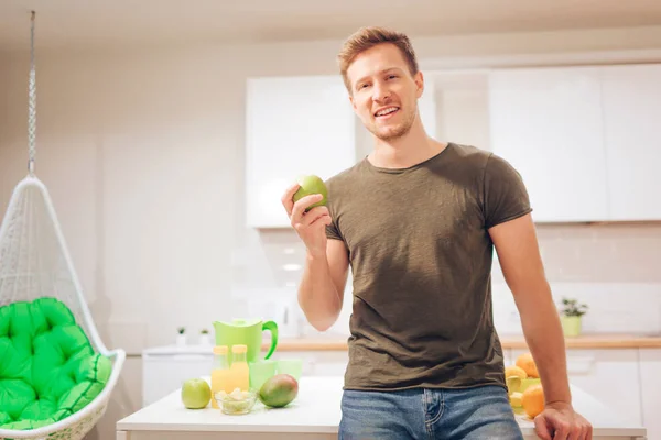 Mladý, usměvavý pohledný muž drží biojablko při řezání čerstvého ovoce v kuchyni. Správná strava. Zdravé jídlo. Vegetariánské jídlo. Detoxikační dieta — Stock fotografie