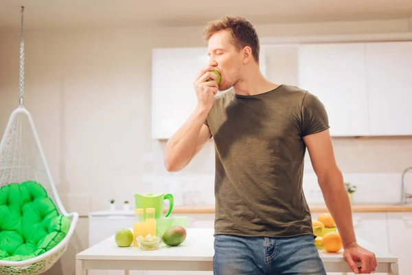 Jovem sorrindo bonito homem morde maçã orgânica enquanto cozinha frutas frescas na cozinha. Alimentação saudável. Refeição vegetariana. Desintoxicação da dieta — Fotografia de Stock