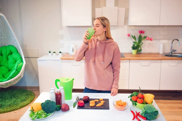 Vegan belle femme blonde boit de l'eau de désintoxication tout en cuisinant des légumes crus dans la cuisine. Alimentation crue. Nourriture végétarienne. Une alimentation saine — Photo