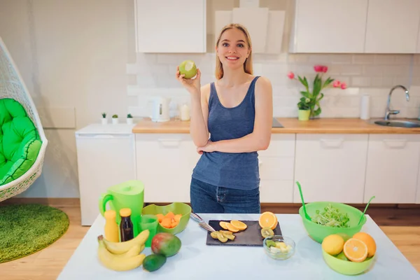 Jeune femme blonde souriante tient la pomme verte mordue tout en cuisinant des fruits frais dans la cuisine. Une saine alimentation. Repas végétarien. Désintoxication alimentaire — Photo