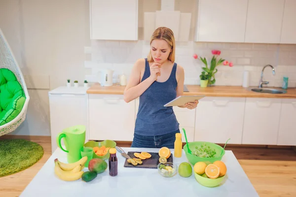 Vegán gyönyörű szőke nő keres egy recept a tabletta bio gyümölcsök a konyhában főzés. Egészséges étel. Detox étrend — Stock Fotó