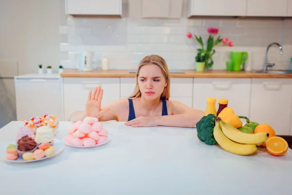 Молодая серьезная женщина в синей футболке выбирает между свежими овощами и сладостями на кухне. Выбор между здоровой и нездоровой пищей. Диета. Диета. Здоровое питание — стоковое фото
