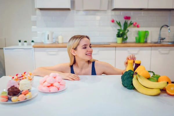 Νεαρή γυναίκα χαμογελαστή στο μπλε T-shirt επιλέγοντας ανάμεσα σε λαχανικά φρέσκα φρούτα ή γλυκά στην κουζίνα. Επιλογή μεταξύ των υγιών και μη υγιών τροφίμων. Να κάνει δίαιτα. Δίαιτα. Υγιεινά τρόφιμα — Φωτογραφία Αρχείου