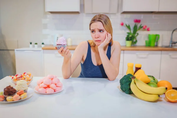 Giovane donna triste con dolce torta in una mano scegliendo tra cibo sano e malsano in cucina. Scelta difficile tra frutta fresca, verdura o dolci. Dieta. Dieta. Cibo sano — Foto Stock