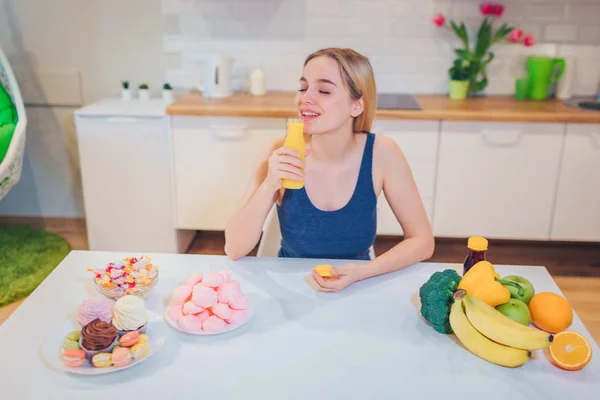 Jovem mulher sorrindo bebe água de desintoxicação ao escolher entre alimentos saudáveis e não saudáveis na cozinha. Escolha difícil entre frutas frescas ou doces. A fazer dieta. Dieta. Alimentos saudáveis — Fotografia de Stock