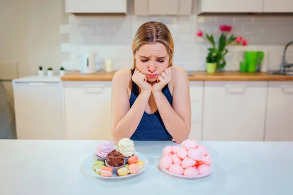 Борьба за диету. Молодая грустная женщина в голубой футболке выбирает между свежими овощами и сладостями, глядя на них на кухне. Здоровое и нездоровое питание. Диета — стоковое фото