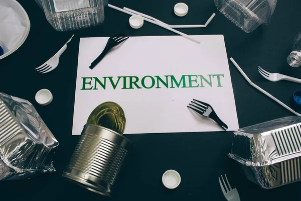 Plastic pollution concept. Paper with word Environment near single-use metal cans, foil containers, straws, broken forks, lids on dark background, top view. Single-use plastic. New rules to reduce — Stock Photo, Image