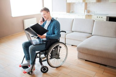 Tekerlekli sandalyede özel ihtiyaçları olan kişi. Genç öğrenci açık kitap bakıyor ve telefonda konuşuyor. Odada tek başına. Gün ışığı.