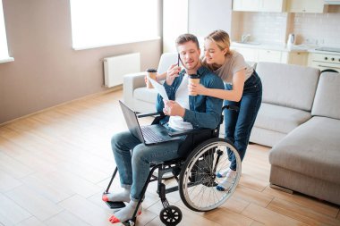 Engelli genç adam tekerlekli sandalyede oturuyor ve arkasına bakıyor. Kadın arkada duruyor ve elinde bir fincan kağıt kahve tutuyor. Erkeğe doğru eğilip gülümse..