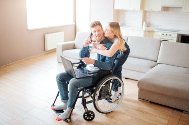 Özel ihtiyaçları olan genç bir adam ve güzel bir kadın. Tekerlekli sandalyede oturuyor ve dizüstü bilgisayarını tutuyor. O arkada duruyor ve ona yaslanıyor. Çift odada birlikte..