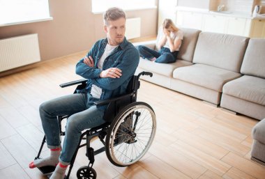 Kapsayıcı ve engelli genç adam tekerlekli sandalyenin önünde oturuyor. Elleri çapraz ve üzgün. Genç bayan kanepede oturup ağlar. Duygusal hastalık ve stres.