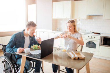 Young man with inclusiveness and special needs eating salad in kitchen. Sit on wheelchair and studying. Young woman sit besides and break eggs. Working together. clipart