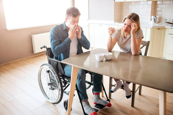 Sick young man with inclusiveness sneezing with healthy woman at table. Sick people in kitchen. Headache and pain. Person with special needs.