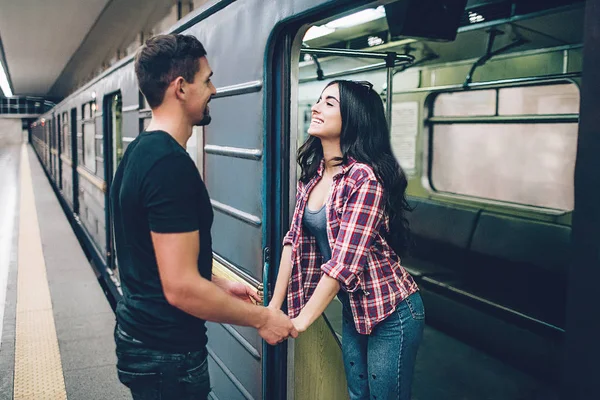 Young man and woman use underground. Couple in subway. Young brunette stand in underground carriage and smile. She hold mans hands. Guy stand on platform. Love stiry. Cheerful. — Stock Photo, Image