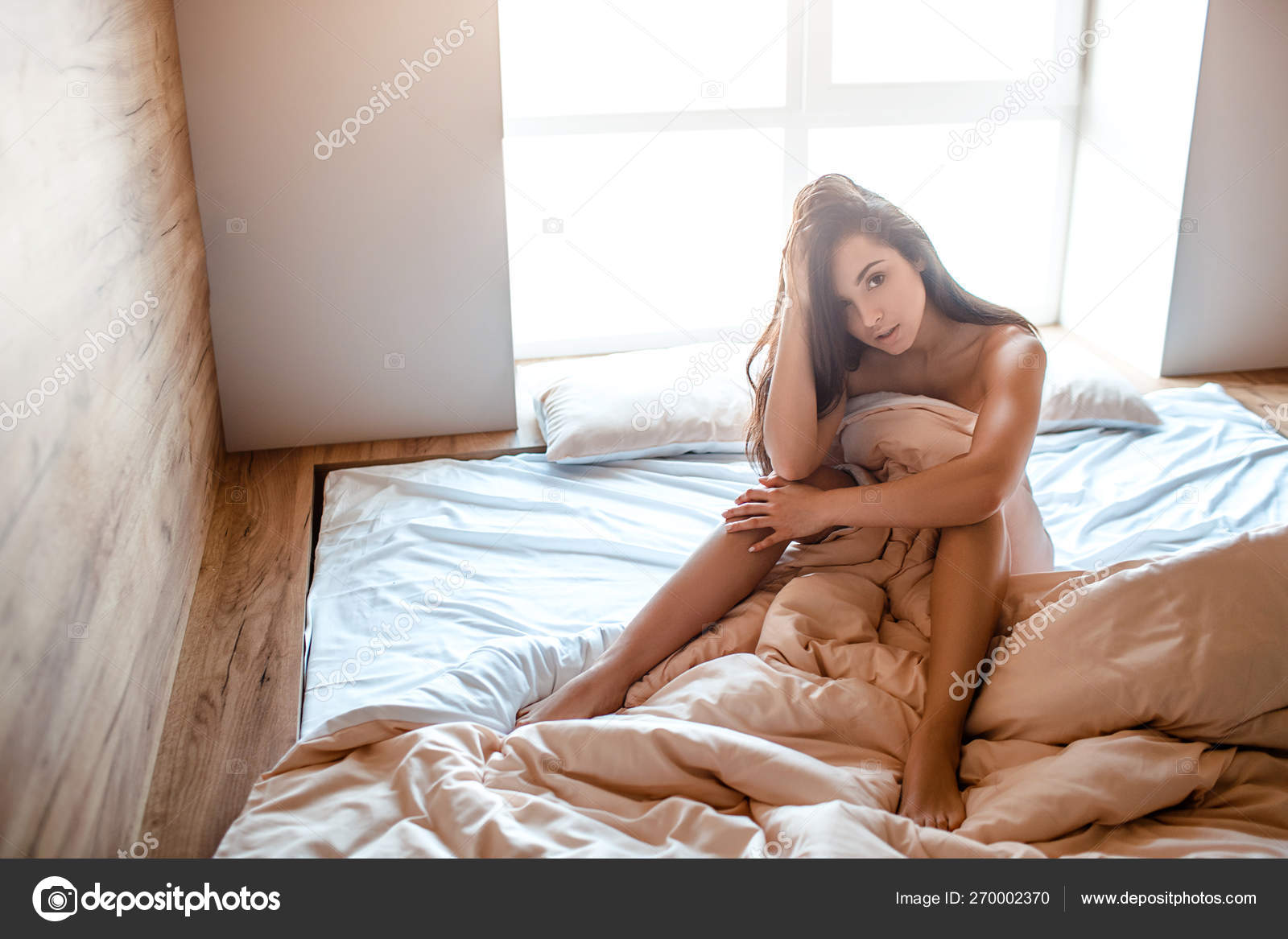 Seductive young naked dark-haired woman on bed in morning. Sexy beautiful model posing on camera. Body covered with blanket. Concentrated. Alone in room