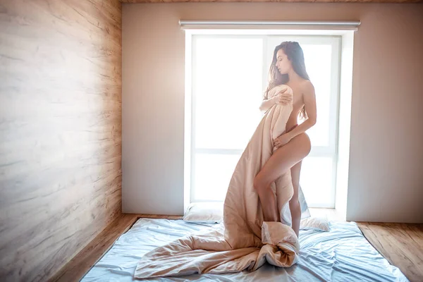 Seductora joven mujer morena desnuda en la cama por la mañana. Hermoso modelo sexy cuerpo de la cubierta con blanco. De pie en la cama. Solo en la habitación . —  Fotos de Stock