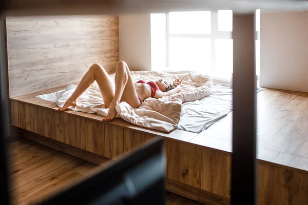 Verführerische junge nackte dunkelhaarige Frau am Morgen im Bett. schlanke heiße Modell allein liegend. gut gebaute schöne Beine. Frau trägt rote Dessous. Masturbation. Sinnliches Bild. — Stockfoto