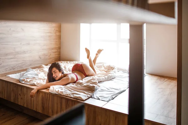 Verführerische junge nackte dunkelhaarige Frau am Morgen im Bett. glücklich fröhliches Model auf dem Bauch liegend und lächelnd. Beine hoch. Tageslicht. — Stockfoto
