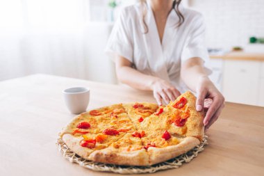 Mutfakta beyaz sabahlıklı kadının kesişme görünümü. Bir parça pizza alıyorum. Küçük bir dilim. Genç hizmetçi kaygısız bir hayat yaşıyor.