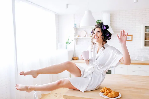 Jeune femme de ménage positive assise sur la table et posant à la caméra. Rire à voix haute. Vague avec la main. Croissants sur assiette. Jeune femme négligente. Curleurs dans les cheveux. Robe de chambre blanche . — Photo