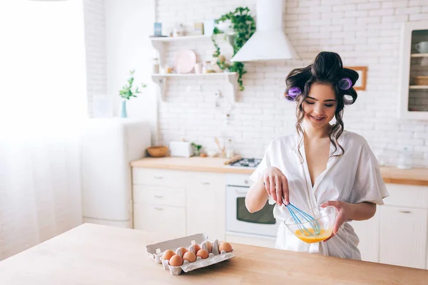 Jeune belle femme de ménage cuisine dans la cuisine. Mélanger les œufs dans un bol en verre. Regarde en bas et souris. Curleurs dans les cheveux. Seul dans la cuisine. Lumière du jour. Porter une robe de chambre blanche . — Photo