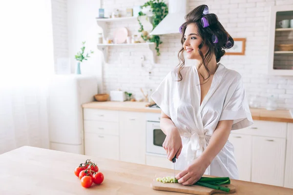 Alegre mujer joven positiva de pie en la cocina y mirar a la ventana. Corte la cebolla verde en el escritorio. La ama de llaves usa bata blanca. Solo en la cocina. Pimienta roja en el lado izquierdo . — Foto de Stock