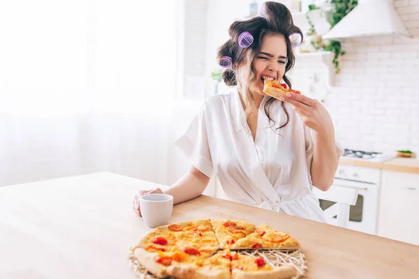 空腹の若い主婦は台所に座ってピザを食べます。食べ物のスライスを噛む。部屋に一人で家政婦は仕事のない生活を楽しむ。カップを手に持つ. — ストック写真